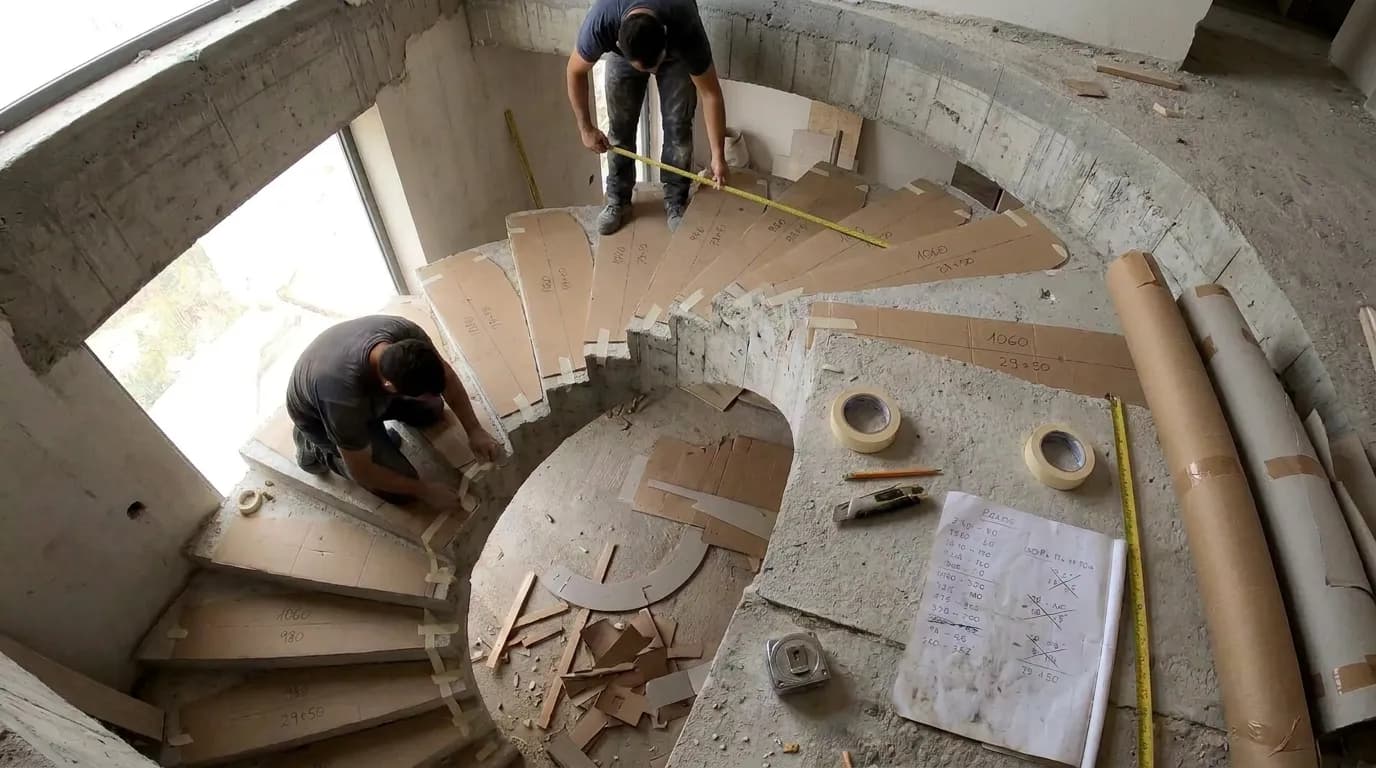 A bare cement curved staircase with a cardboard template cut and taped to each tread, offcuts, tape and handwritten notes, two workers measuring by hand